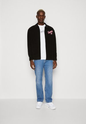 Schwarze Fleecejacke mit roter und weißer Stickerei, getragen über einem weißen T-Shirt, kombiniert mit blauen Jeans und weißen Sneakers.