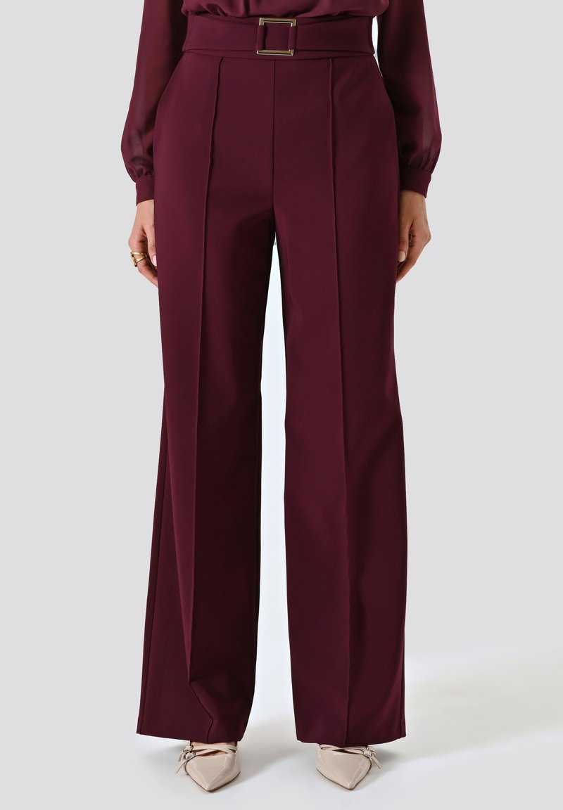 Pantalons larges bordeaux avec une taille ceinturée, des plis sur le devant et un tissu lisse. Associés à des chaussures beiges à bout pointu.