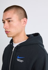 Ung mand med kort hår iført en sort zip-up hættetrøje med et blåt Nike-logo og teksten "ATHLETIC DEPT JUST DO IT." på brystet.