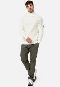 Pull à col roulé en tricot crème avec une texture côtelée, associé à un pantalon olive et des baskets blanches avec des accents gris et jaunes.