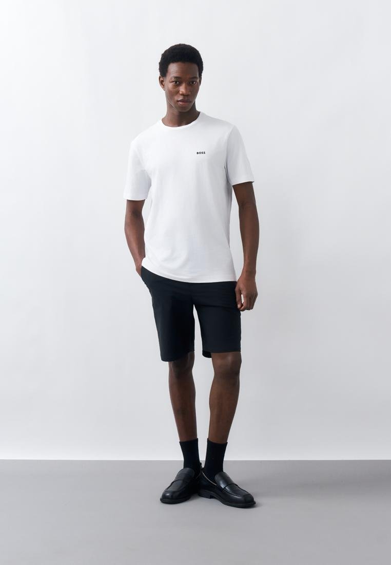 Homme portant un t-shirt blanc "BOSS", un short noir, des chaussettes noires et des mocassins noirs, debout avec une main dans la poche devant un fond uni.