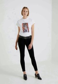 Bijela pamučna majica s grafikom i natpisom "BAG IT UP" sprijeda, uparena s crnim uskim trapericama i crnim cipelama na špic.