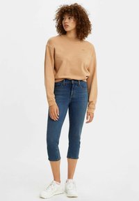 Bruine sweatshirt met geribbelde boorden, gecombineerd met cropped blauwe jeans. Model draagt witte sneakers en toont een casual, relaxte uitstraling.