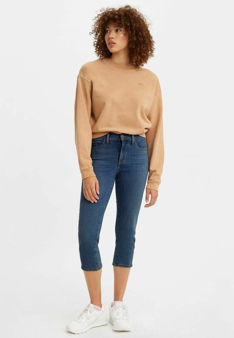Bruine sweatshirt met geribbelde boorden, gecombineerd met cropped blauwe jeans. Model draagt witte sneakers en toont een casual, relaxte uitstraling.