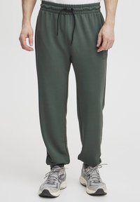 Zelené športové joggingové nohavice s elastickým pásom, nastaviteľnou šnúrkou, bočnými vreckami a zúženými nohavicami. Nosené s šedými teniskami.
