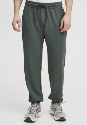 Pantalons de jogging sportifs verts avec une ceinture élastique, un cordon de serrage ajustable, des poches latérales et des jambes fuselées. Portés avec des baskets grises.