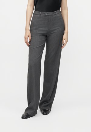 Personne portant un pantalon large gris foncé et des chaussures noires, debout sur un fond blanc uni.