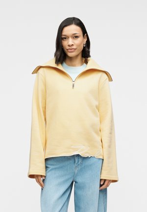 Sweatshirt jaune à col montant, fermeture éclair quart de devant et manches longues. Présente un logo brodé blanc sur l'ourlet.