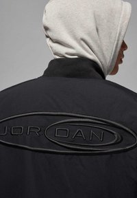 Zwarte jas met opstaande "JORDAN" logo en ronde accenten, gedragen met een grijze hoodie eronder. Gladde stofstructuur.