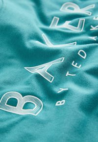 Gros plan sur un tissu bleu sarcelle avec des lettres blanches en contour formant partiellement le mot « BAKER » et un texte plus petit en dessous. Le tissu est texturé et légèrement froissé.