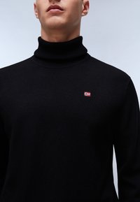 Zwarte coltrui van gebreide stof, met een klein geborduurd vlagdetail op de borst en een gladde textuur.