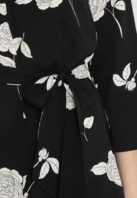 Vestido negro con patrones de rosas blancas, con cintura ajustada y mangas de tres cuartos. La tela parece suave con un acabado ligeramente texturizado.