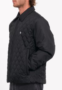 Chaqueta negra acolchada con cuello alzado, cierre frontal de cremallera y bolsillos laterales. El tejido parece suave con un patrón de diamante texturizado.