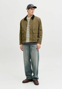 Jeune homme portant une veste vert olive, un pull rayé beige, un jean bleu ample, des chaussures marron et une casquette noire, debout devant un fond blanc.