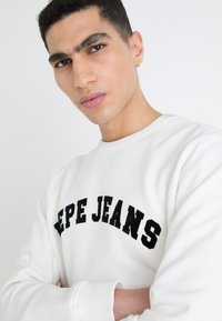 Witte sweater met zwarte "PEPE JEANS" letters over de borst. Gladde stof, ronde hals en geribbelde manchetten. Close-up van het gezicht van het model.