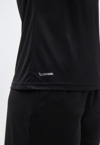 Μαύρο αθλητικό πουκάμισο με τελείωμα πάνω από αντίστοιχα σορτς, με μικρό λευκό λογότυπο "Climacool" κοντά στην κάτω άκρη στην πλευρά.