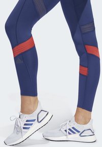 Granatowe legginsy zdobione są wyrazistymi czerwono-złotymi paskami, zestawione z białymi butami sportowymi z niebieskimi paskami i teksturowanym dzianym wierzchem.