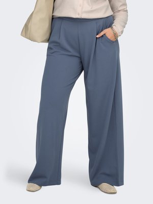 Femme portant un pantalon large bleu, un haut beige, avec un sac beige, la main dans la poche et des chaussures blanches tissées visibles.