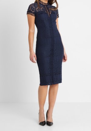 Laivastonsininen pitsimekko, jossa on lyhyet hihat, korkeakauluksinen, kukkakuvioitu ja vartalonmyötäinen siluetti, ulottuen juuri polven alapuolelle.