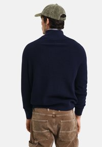 Granatowy sweter z prążkowanej dzianiny z wysokim kołnierzem, noszony z brązowymi spodniami z korduroy; ma długie rękawy i luźny krój.