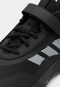 Czarne buty sportowe z teksturowanej siateczki i syntetycznego materiału, płaskimi sznurówkami i zapięciem na rzep. Ozdobione odblaskowymi pasami z boku.