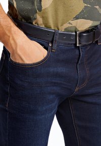 Vaqueros de mezclilla azul oscuro con detalles de costura naranja, que incluyen un cinturón de cuero negro con una hebilla plateada. El bolsillo es parcialmente visible.