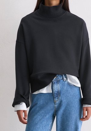 Persoon draagt een zwart cropped coltrui over een gestreept wit shirt met blauwe hoog getailleerde jeans, handen ontspannen langs de zijden.