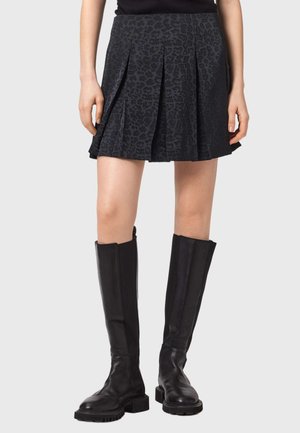 Nainen, joka pukeutunut mustaan, liehuvaan minihameeseen, jossa on hillitty leopardikuvio ja mustat polven yläpuolelle ulottuvat nahkasaappaat.