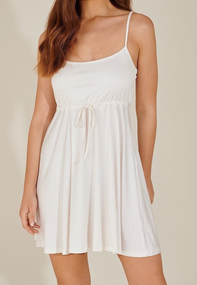 Femme portant une robe blanche sans manches avec de fines bretelles et un détail noué à la taille, debout devant un fond clair uni.