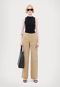 Zwarte geribbelde mouwloze top, beige wijde broek met stikselaccenten, en zwarte sandalen. Model houdt een zwarte handtas vast.