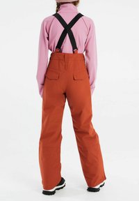 Orangene wasserdichte Skihosen mit verstellbaren schwarzen Trägern, weitem Bein und Gesäßtaschen, kombiniert mit einem rosa Langarmshirt.