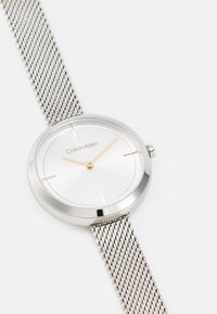 Reloj de pulsera de malla plateada con una cara redonda, esfera blanca, agujas doradas y un diseño minimalista. Presenta el nombre de la marca "Calvin Klein" debajo de las 12 en punto.
