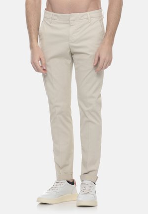 Uomo che indossa pantaloni chino beige slim fit con orli arrotolati e sneakers bianche, in piedi contro uno sfondo chiaro e semplice.