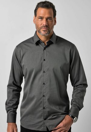 Homme aux cheveux courts et foncés avec une barbe, portant une chemise à manches longues gris foncé boutonnée et une montre-bracelet à la main gauche.