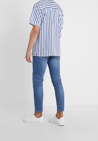 Hombre vestido con una camisa de manga corta con rayas verticales azules y blancas, jeans ajustados azules y zapatillas blancas, de pie con la espalda hacia la cámara.