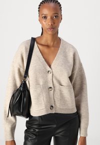 Beige wolmix cardigan met knoopsluiting, geribbelde zoom en twee voorzakken. Gepaard met zwarte leren broek en een zwarte tas.
