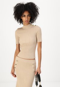 Beige ribbestrikket topp med høy hals og korte ermer, med knappdetaljer på skulderen, kombinert med et matchende skjørt.