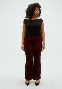 Fekete váll nélküli felső, bordó, széles szárú, bársonyos textúrájú nadrág, fekete magassarkú cipővel párosítva, háttér előtt álló nő.