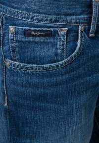 Jeans de mezclilla azul con un bolsillo delantero, costuras detalladas y una etiqueta de "Pepe Jeans". La tela tiene una textura suave con un leve desgastado.