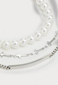Trois bracelets : un fil de perles blanches, une chaîne en argent avec des pendentifs croix, et un bracelet bar en argent élégant avec un bord texturé.