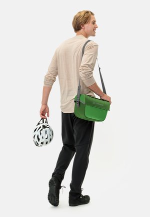 Borsa a tracolla verde realizzata in materiale resistente, con chiusura a pattina e tracolla regolabile, tenuta da una persona con un casco da bicicletta.