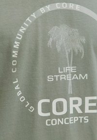 Camiseta de algodón verde oliva con texto impreso en blanco y un gráfico de palmera, diseño circular con frases como "CORE" y "COMUNIDAD GLOBAL."