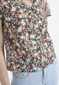 Blouse à motif floral avec un décolleté en V, des manches courtes et des boutons à l'avant. Le tissu semble léger et coloré sur un fond sombre.