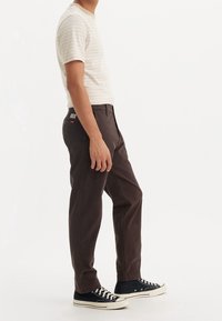 Homme portant un pantalon marron, des baskets montantes noires et une chemise à manches courtes beige rayée, debout de profil sur un fond blanc.