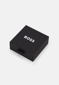 Juodas stačiakampis dėžutė su matinės apdailos ir tekstūruotu rankenėle, viršuje esantis baltas logotipas "BOSS".
