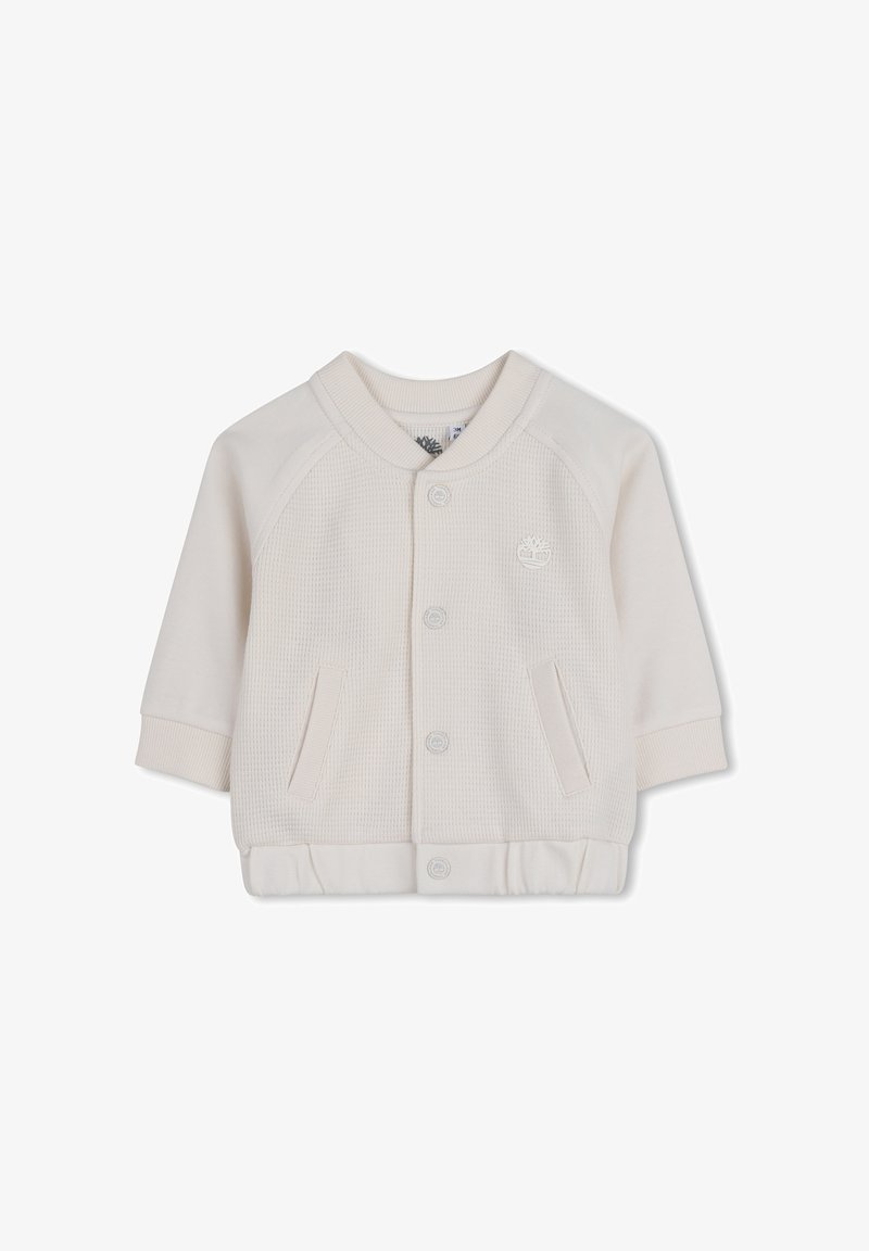 Veste beige clair en tissu texturé, col et poignets côtelés, fermeture à boutons sur le devant, et deux poches latérales. Présente un petit logo sur la poitrine.