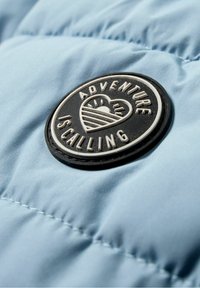 Patch rond noir et blanc sur tissu bleu montrant un cœur avec un lever de soleil et le texte « L'aventure appelle. »
