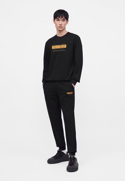 Млад мъж, облечен с черен суитшърт EA7 Emporio Armani и съвпадащи черни спортни панталони с черни маратонки на бял фон.