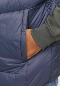 Marineblauer Puffer-Weste mit einem gesteppten Design; sie hat eine große Seitentasche und einen grau umgeschlagenen Ärmel, der in der Nähe sichtbar ist. Glatte, glänzende Textur.