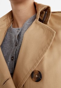 Beige trench coat featuring a notched collar, textured fabric, and a large dark button. Worn over a gray knitted sweater with buttons.